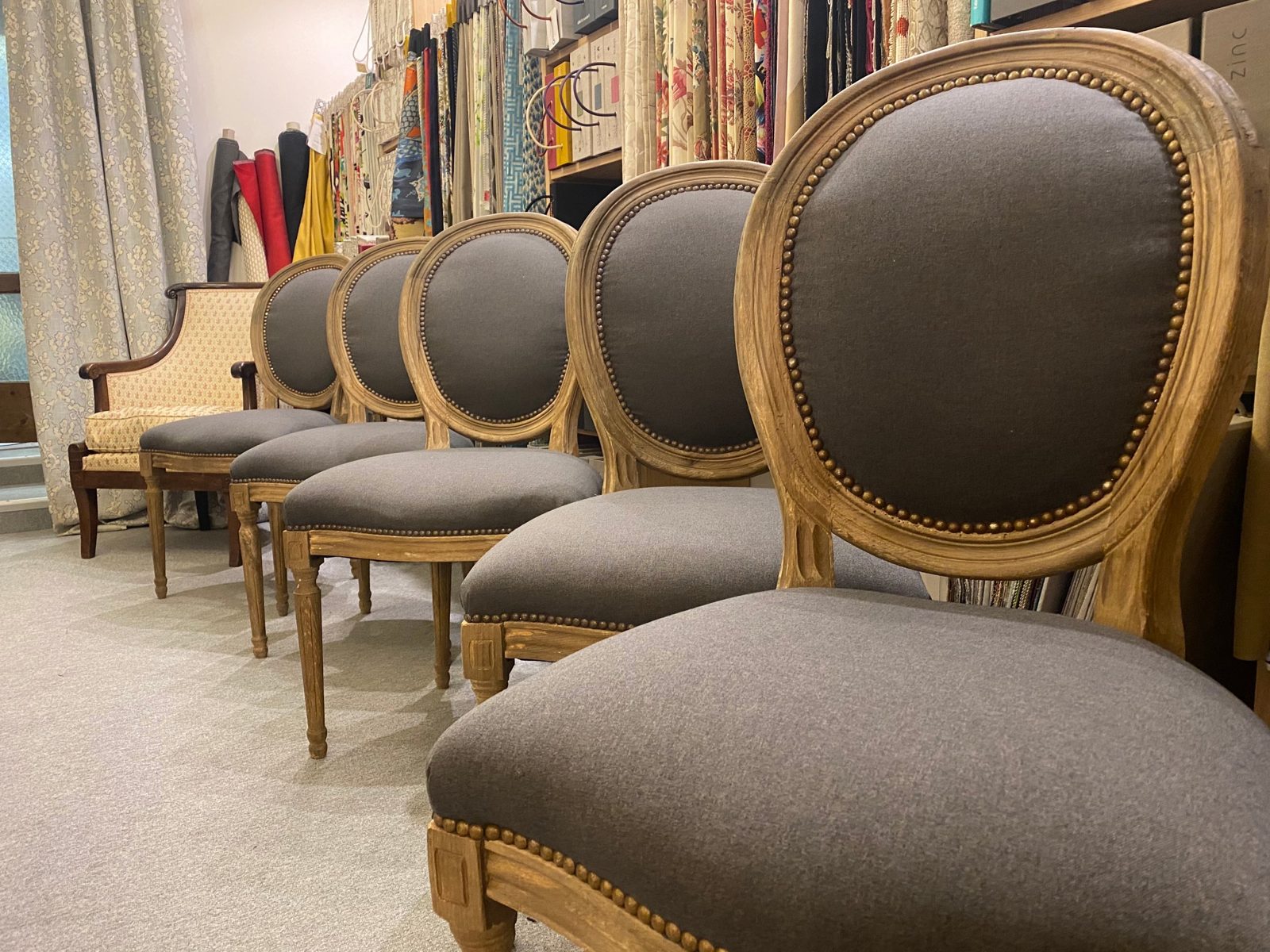 Réfection de chaises : cinq chaises à dossier médaillon alignées, recouvertes d’un tissu gris avec clous de tapissier, dans l’atelier