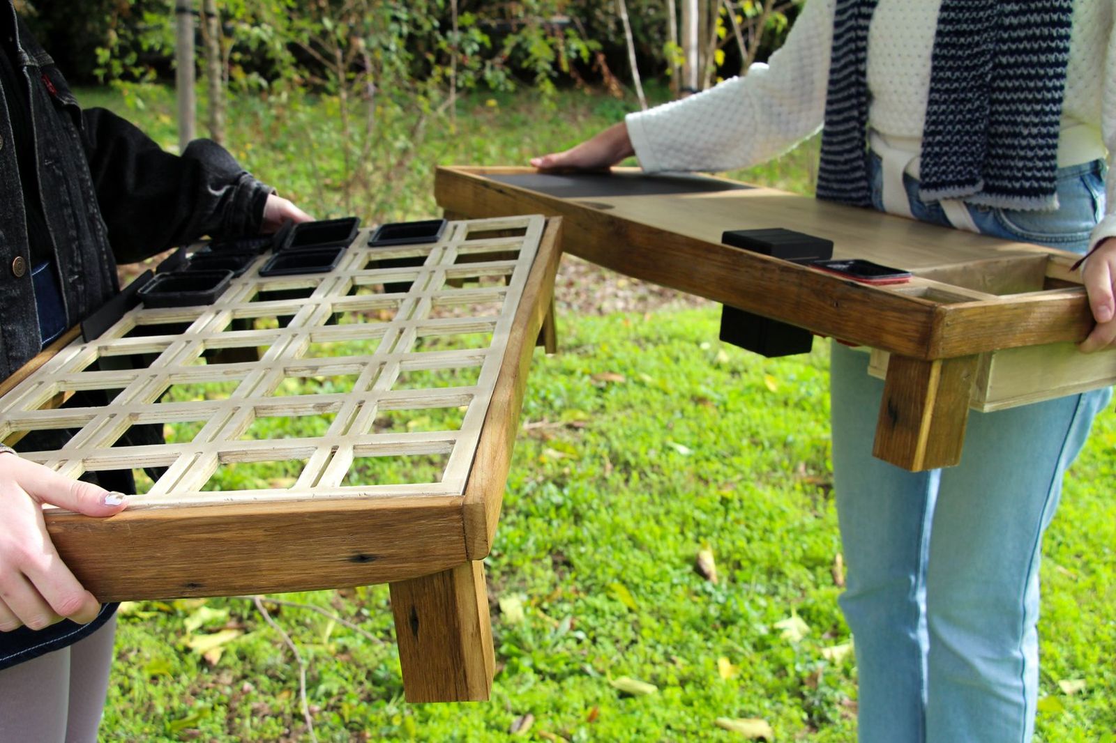 Deux personnes portent chacune un plateau en bois : à gauche le plateau de rangement en quadrillage avec ses pots noirs, à droite la table de travail, dans un jardin