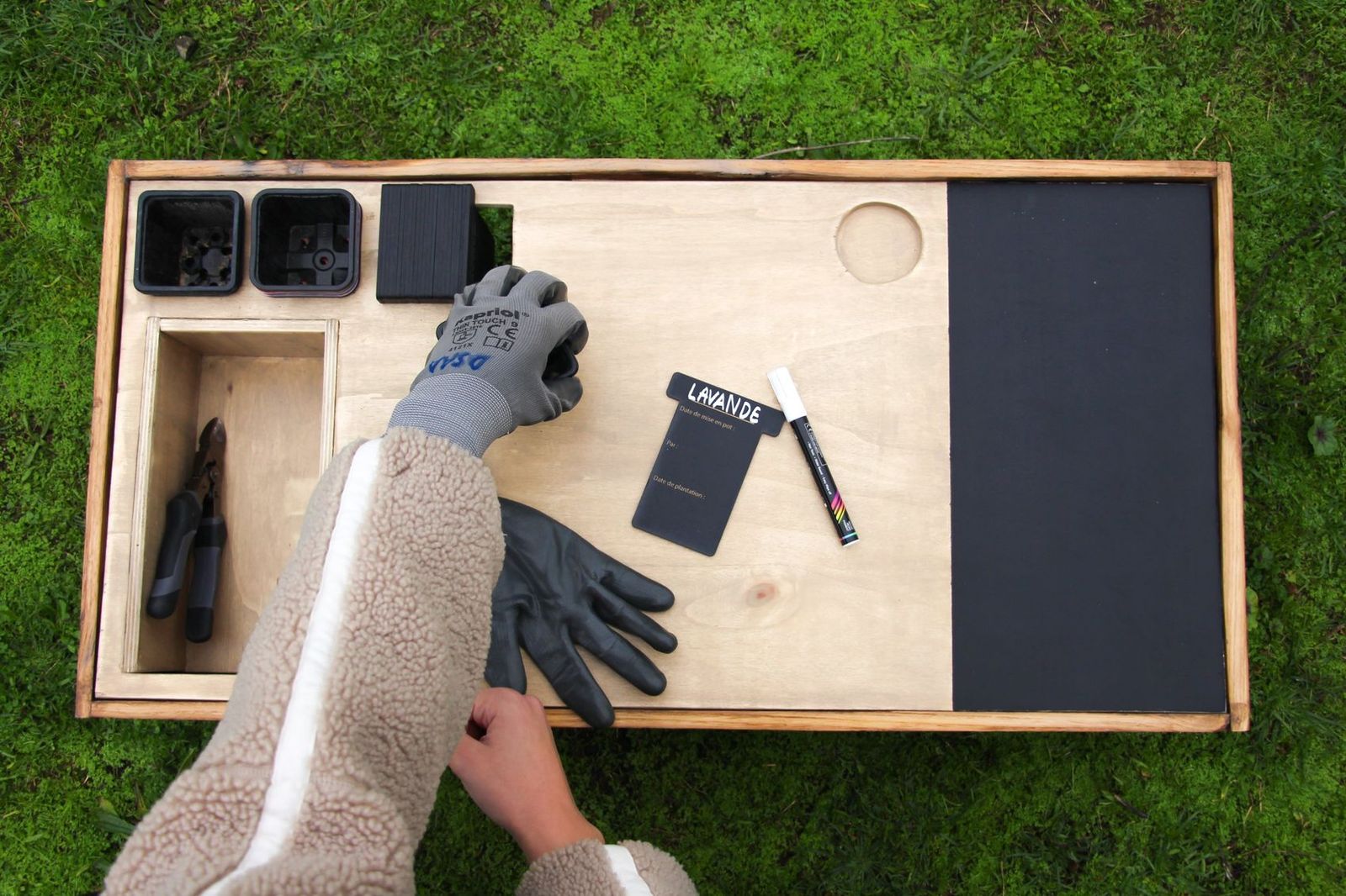 Table de travail vue du dessus : pots, bac à outils avec sécateur, gants, carte « Lavande », feutre blanc et ardoise noire pour les notes