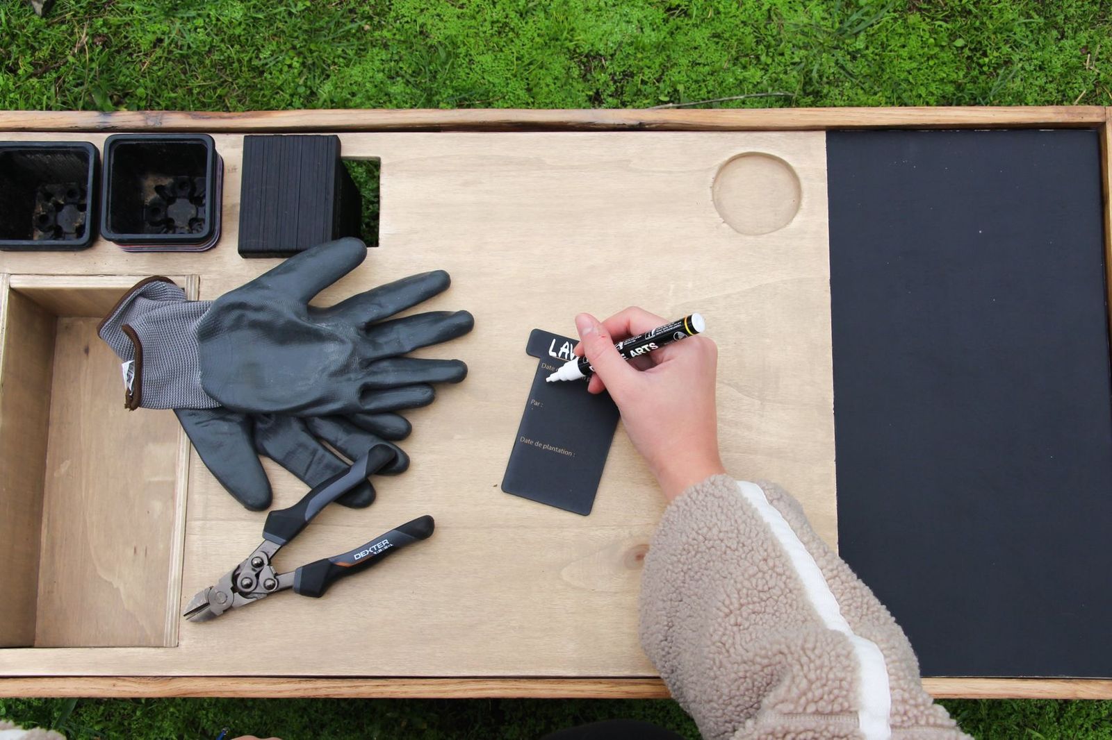 Une main écrit « Lavande » au feutre blanc sur une carte noire posée sur la table de travail, à côté des gants et du sécateur